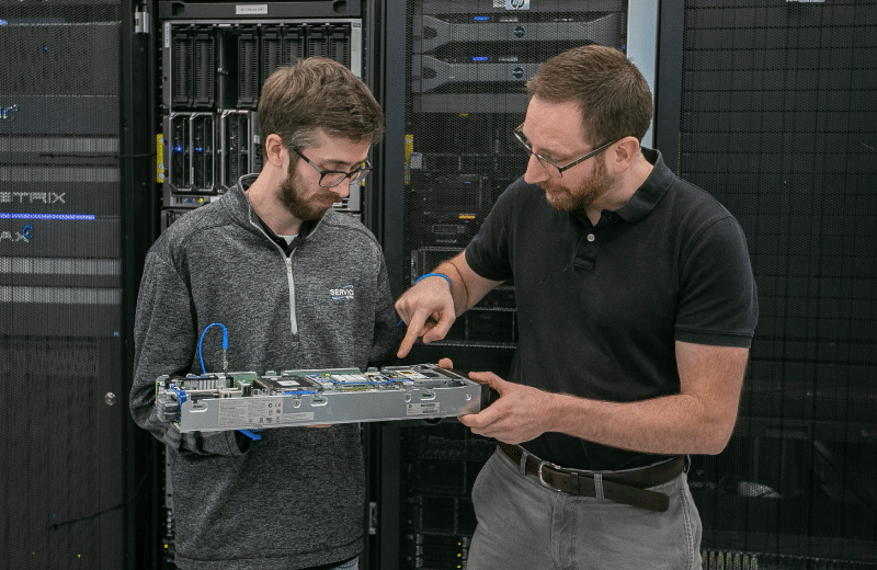 An image of two engineers in a data center. One engineer is holding a piece of equipment as the other engineer points to parts within the equipment