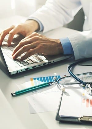 An image of a doctors hands typing on a laptop with some papers a pen and a stethoscope next to the laptop