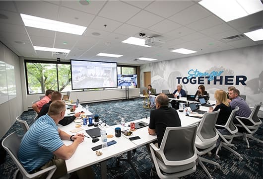 An image of a group gathered for a meeting in a conference room with another group on a video call on the projection screen