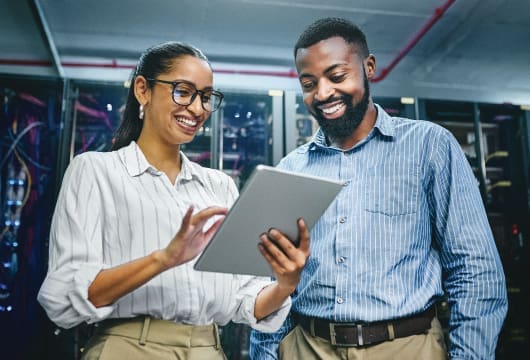 An image of a female and male engineer in a data center. The female engineer is holding a tablet and scrolling it. She is tilting the tablet so the male engineer can see what is on it and they are both smiling.