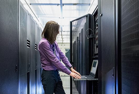 An image of a male engineer with long hair typing on a laptop connected to equipment within a data center