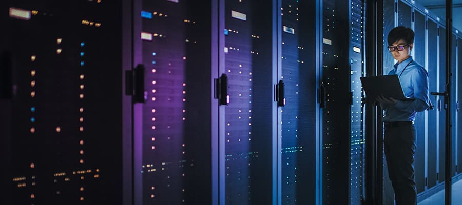 An image of an engineer in a dimly lit data center holding and looking into a laptop