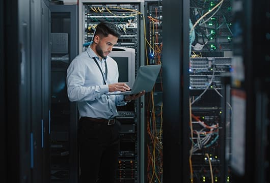 An image of a male engineer holding a laptop standing in front of open equipment racks