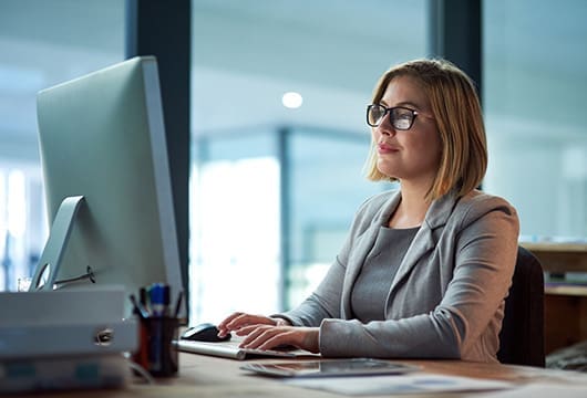 An image of a woman sitting at a desk smiling and typing on their computer