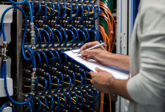 An image of hands holding clipboard and pen in front of equipment with cables