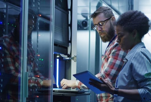 An image of a male engineer typing on a laptop and a female engineer holding a tablet in a data center