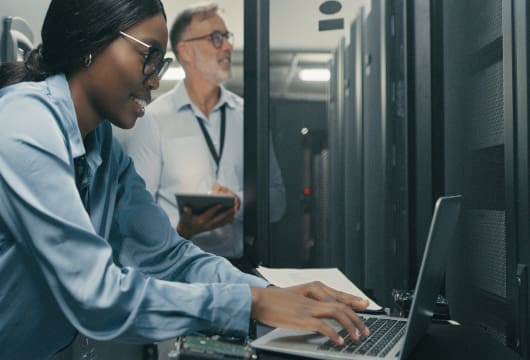 An image of a female engineer smiling and typing on a laptop and a male engineer checking equipment in the background