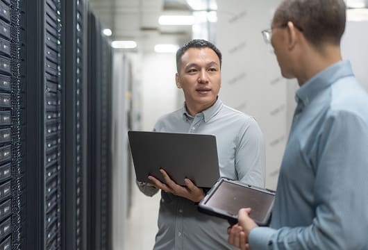 An image of two engineers in a data center, one holding a laptop