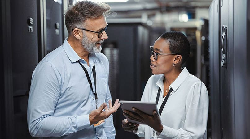 An image of two engineers in a data center. They are smiling and conversing. The female engineer is holding a tablet.