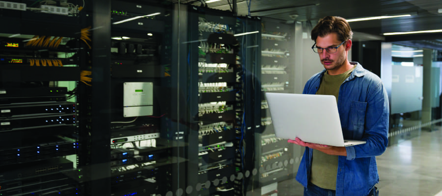 An image of a male engineer holding a laptop standing in front of equipment racks in a data center