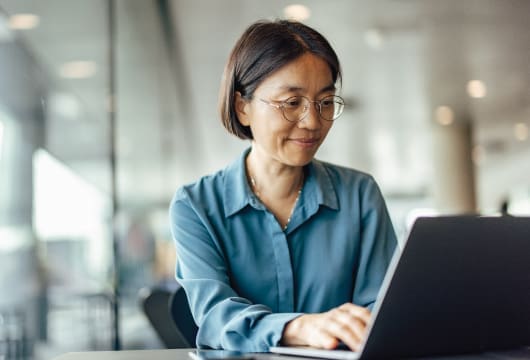 An image of a female professional with round wire glasses smiling sitting at a laptop with hands positioned on keyboard