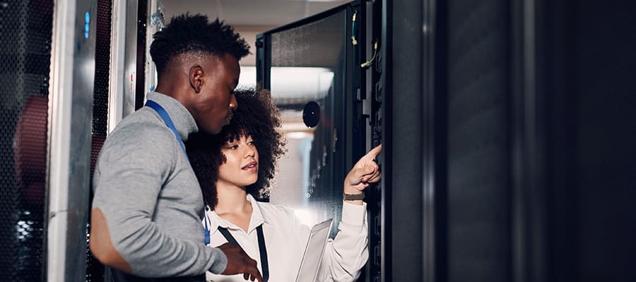 An image of a male and female engineer standing in front of a data center equipment rack. The female engineer is pointing to equipment within the rack.