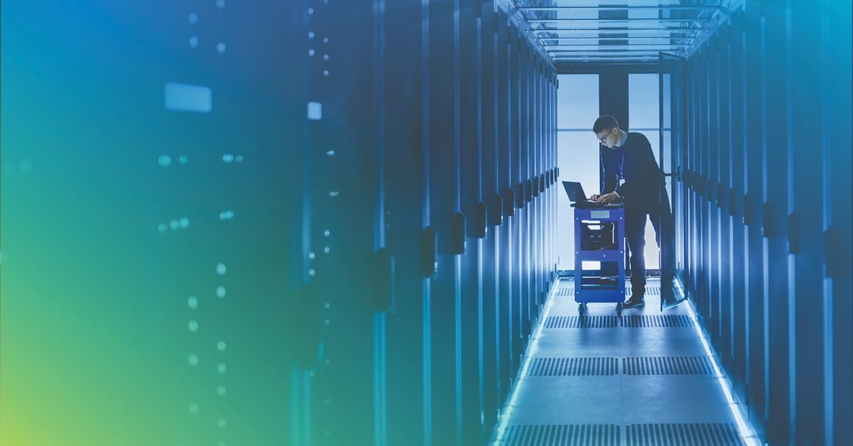 An image of an engineer typing on a laptop on top of a cart in a data center hall surrounded by equipment racks