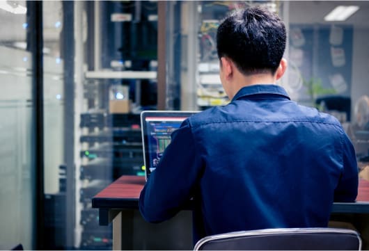 An image from behind a person sitting at a desk on a laptop with data center equipment in the background