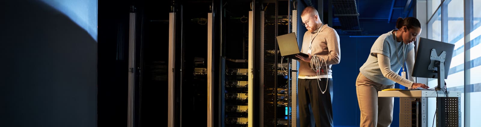 An image taken in a data center of a male engineer with a cable around his arm holding a laptop, a female engineer at a desktop