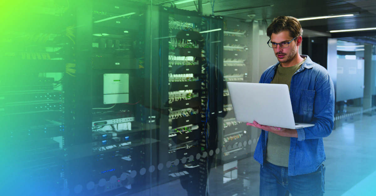 An image of a male engineer holding a laptop standing in front of equipment racks in a data center